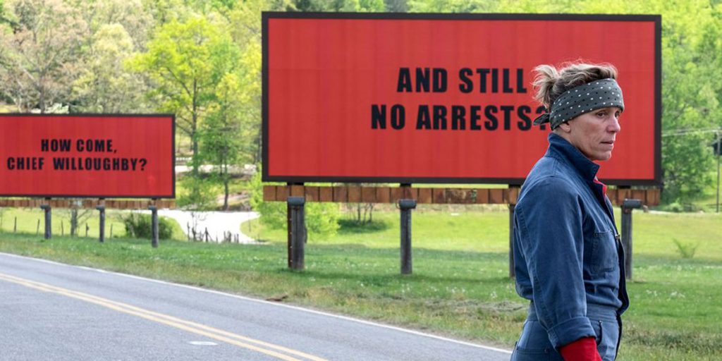 Frances McDormand stands in front of a red billboard that reads "and still no arrests?" in a still from the film Three Billboards Outside Ebbing, Missouri