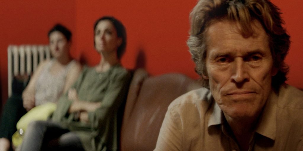 Willem Dafoe looks at the camera in front of a red background in the film Tommaso
