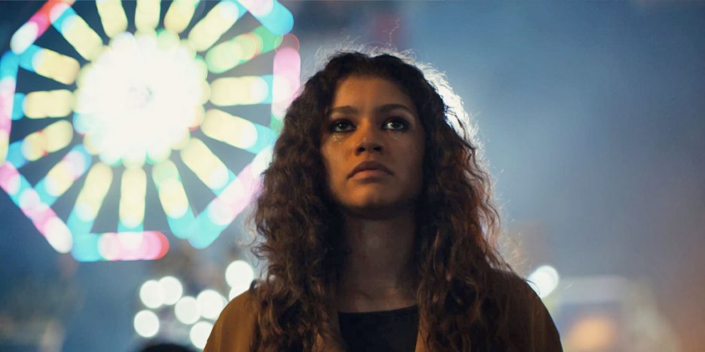 Zendaya, as Rue, stands in front of a ferris wheel at night in the series Euphoria