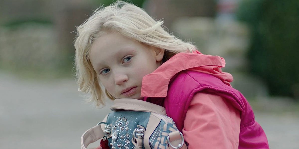 Image is from the film 'System Crasher' (2019). A young girl is a pink jacket holds a jewelled bag to her chest.