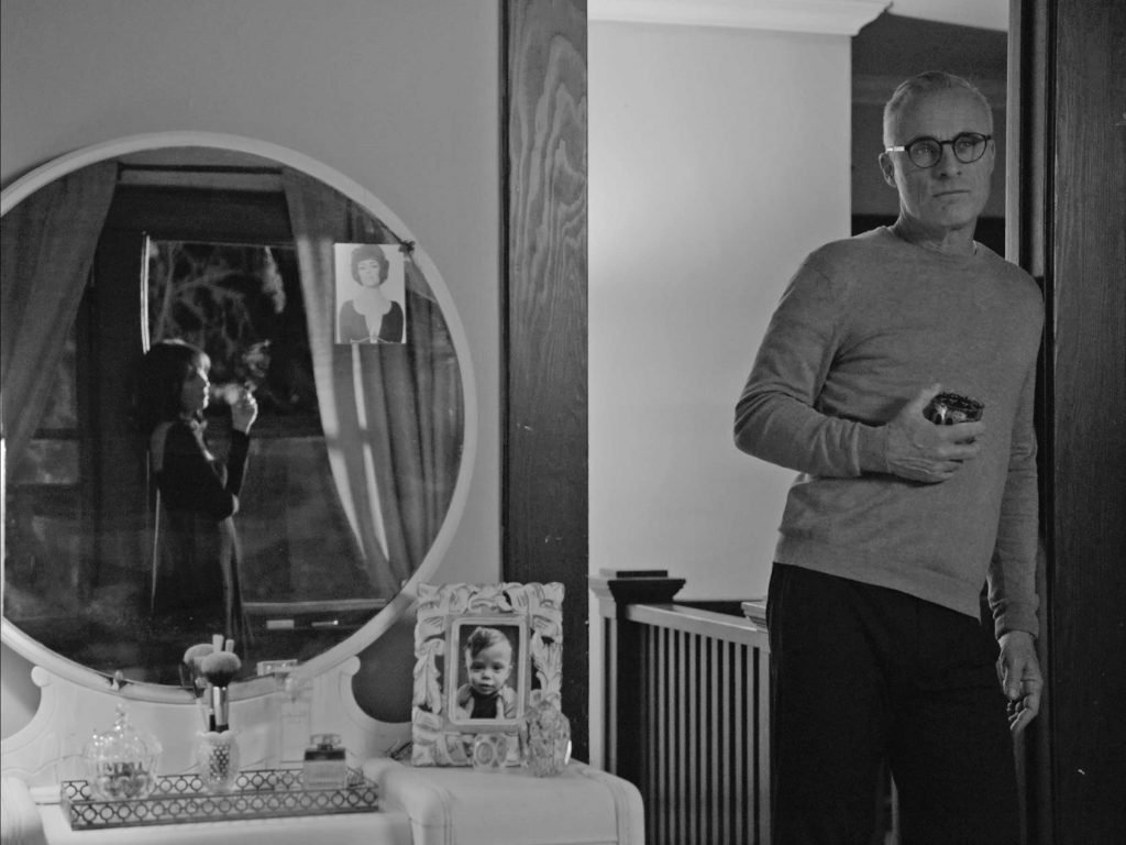 A man stands holding a glass with a mirror next to him, looking at a woman, in a black and white still from the 2021 film "New Year"