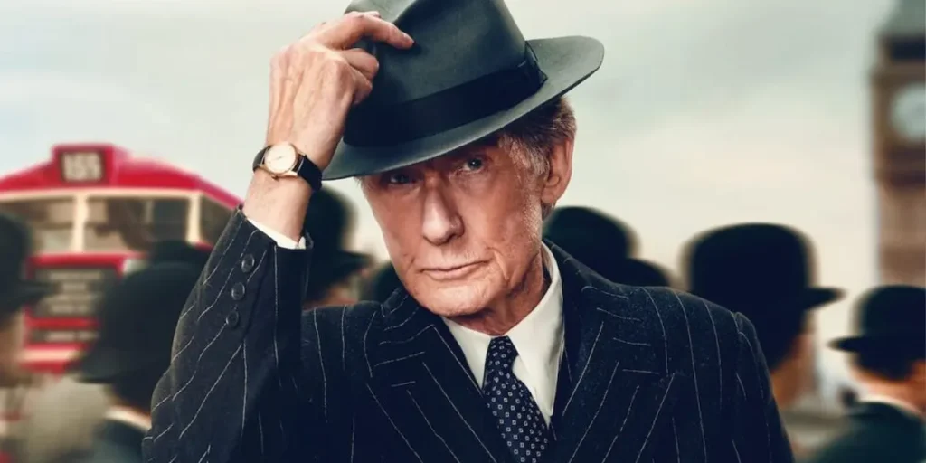 Bill Nighy touches his own hat with a bus behind him in the 2022 film "Living"