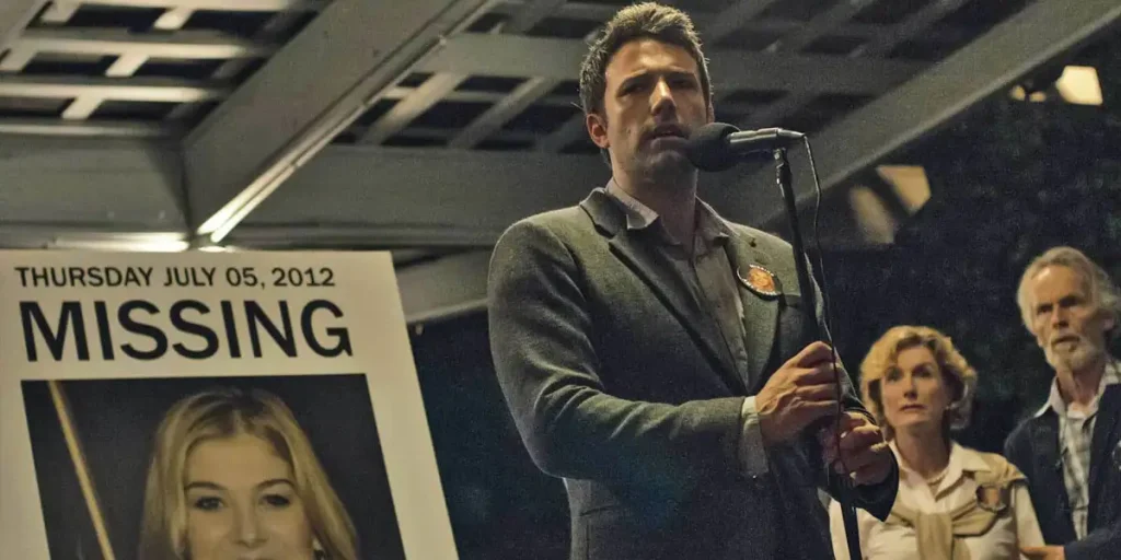 Ben Affleck stands next to a photo that reads "missing" and shows Rosamund Pike's face in a still from the movie Gone Girl