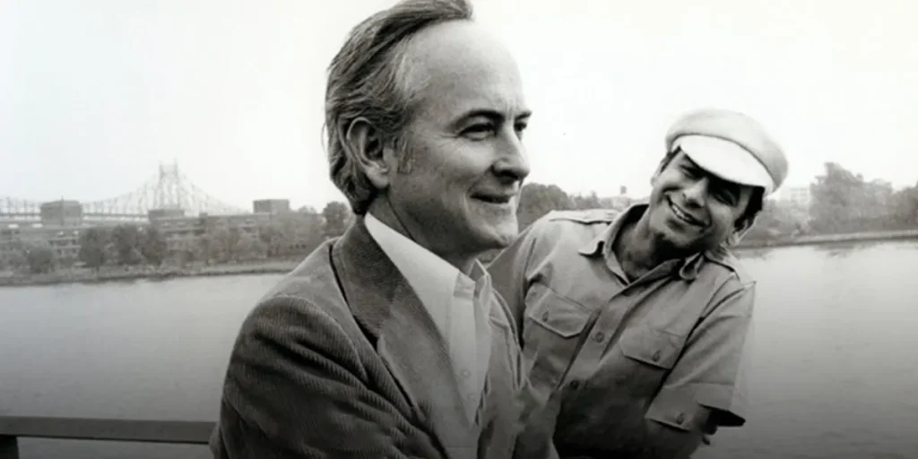 Two men smile by a river in a still from the film Merchant Ivory