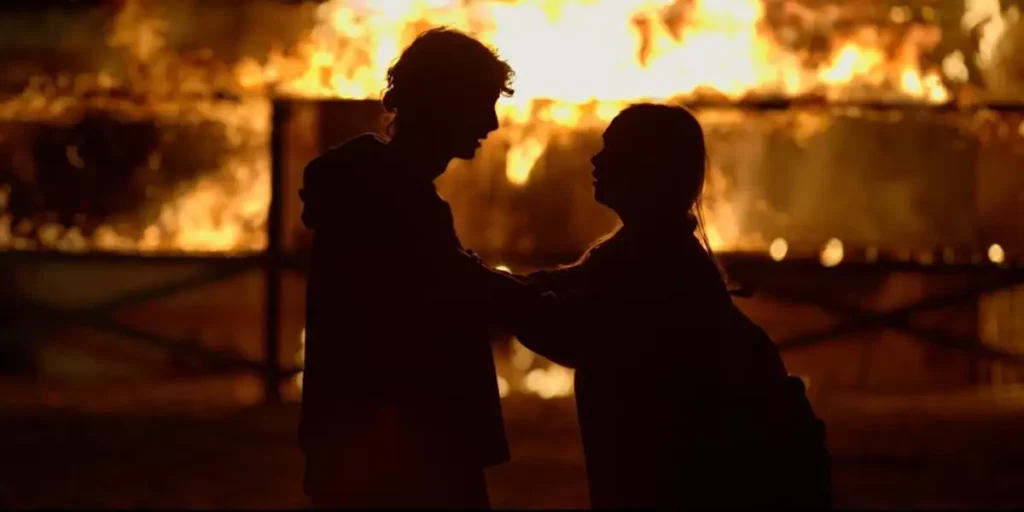 Two characters stand in front of each other, hands on each other's shoulders, with fire burning behind them in the film The Burning Season