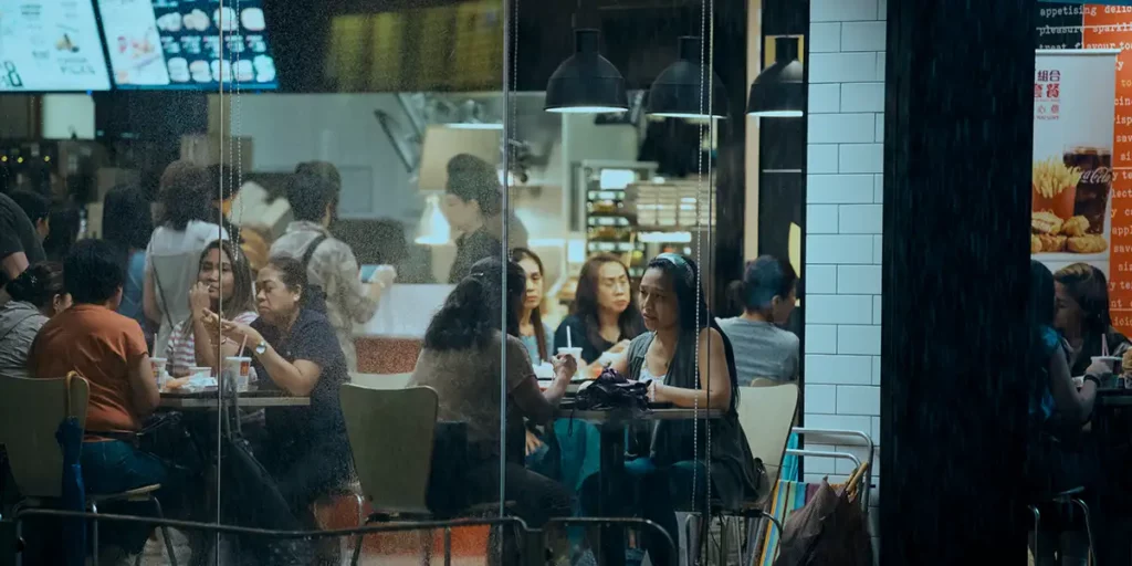 Puri (Amelyn Pardenilla) and Essie (Ruby Ruiz) sit at a table behind glass doors in Episode 5 of Expats