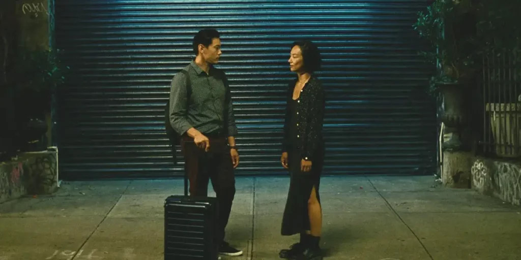Teo Yoo and Greta Lee look at eachother in the middle of the street holding a suitcase