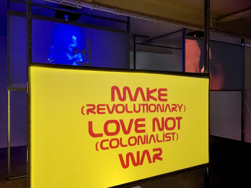 Red writing on a yellow background reads "make (revolutionary) love not (colonialist) war" in a/political's exhibition for Bruce LaBruce's art project-turned-film The Visitor