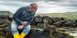 Billy Mail and his otter friend sit next to each other by the beach in the film Molly in Billy & Molly: An Otter Love Story