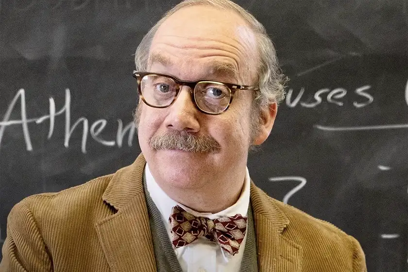 Paul Giamatti stands in front of a blackboard and smiles as Paul Hunham in The Holdovers