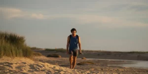 A man walks on the beach holding his sunglasses and hat in the film High Tide