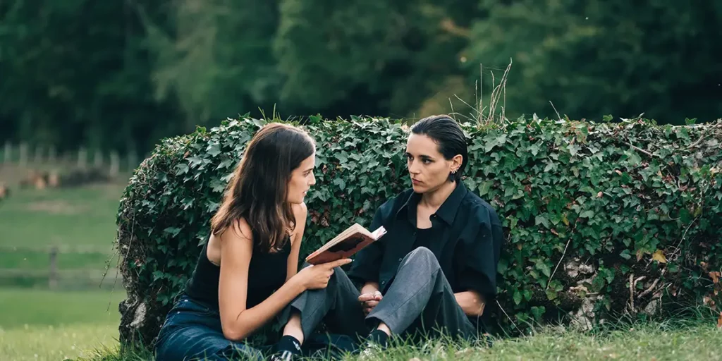 A woman holds a book and reads to another woman while sitting in the park in the 2024 film Split, from BFI Flare