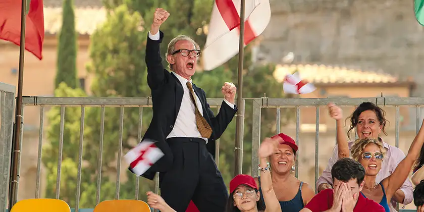 Bill Nighy jumps and shouts in victory with an arm raised during a football match in Netflix film The Beautiful Game