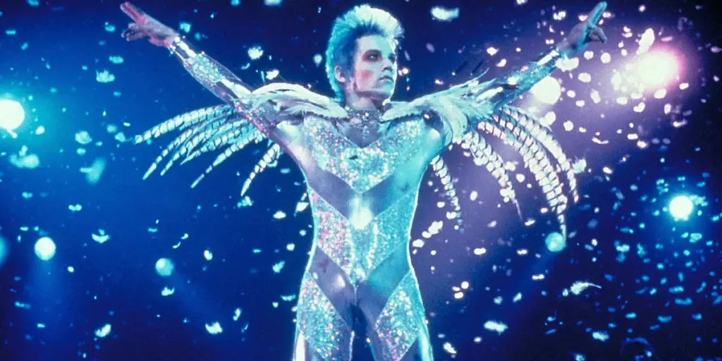 A man dressed in a shiny white jumpsuit reminiscent of Ziggy Stardust has their arms open during a concert in the film Velvet Goldmine