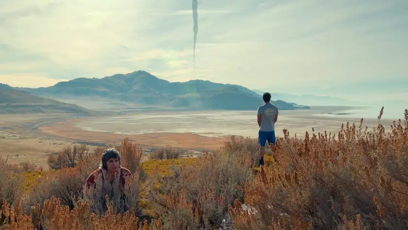 A woman crouches on the ground while a man faces the other way standing up, facing the mountains in a field in the film We're All Gonna Die