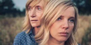 Two women stand one behind the other, facing the camera, looking sad in the film Woman Of...