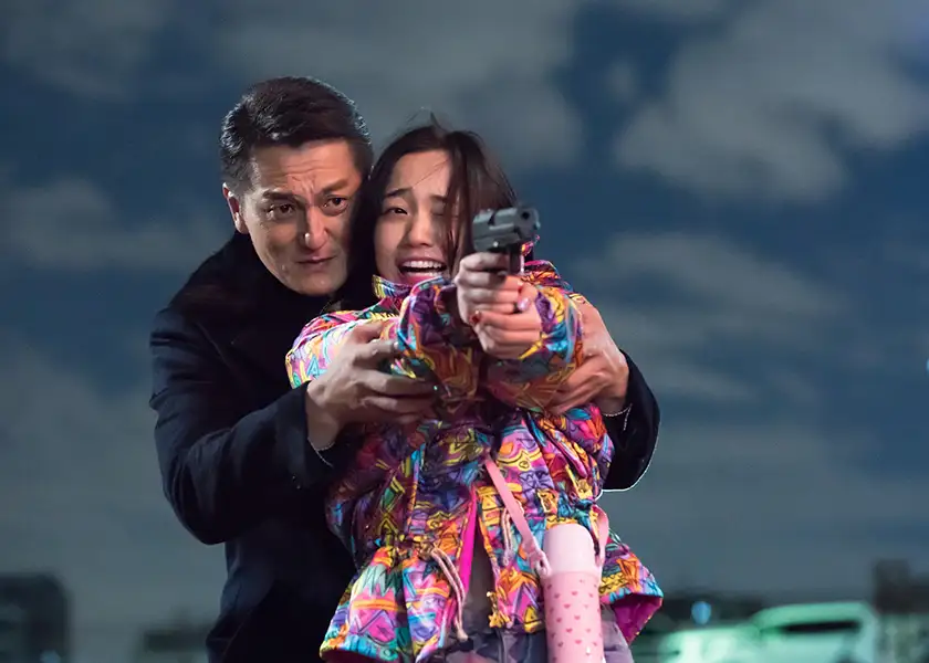 A man stands behind a girl holding a gun and wearing a pink jacket in the film Baby Assassins (2021)