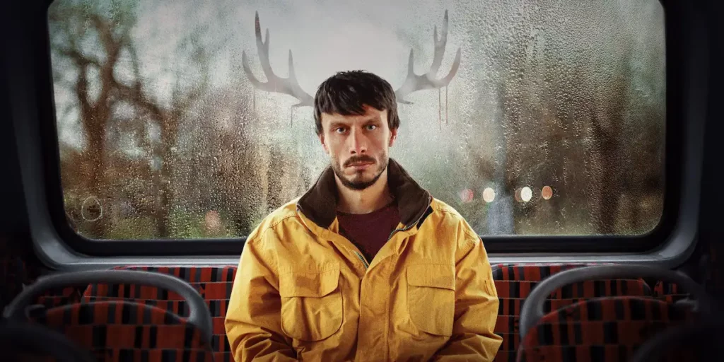 Richard Gadd as Donny wears a yellow hacket and has reindeer antlers drawn on a window behind him in a still from the Netflix series Baby Reindeer