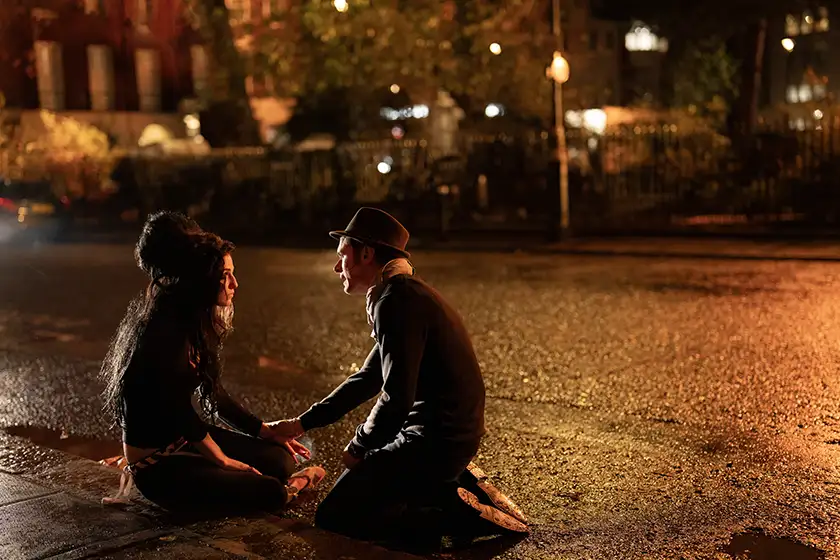 Marisa Abela as Amy Winehouse and Jack O’Connell as Blake Fielder-Civil sit on the ground in a road where it's just rained in the film Back to Black