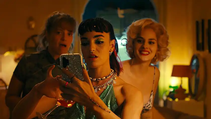 Three women are dressed for a party and look at a glittery iphone screen in the film The Balconettes, one of the 20 films to watch at the 2024 Cannes film festival according to Loud and Clear Reviews