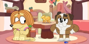 Two of the supporting characters of the series Bluey - a yellow dog with long hair and a pitbull - stand next to a table with a statue on it in an episode of the show