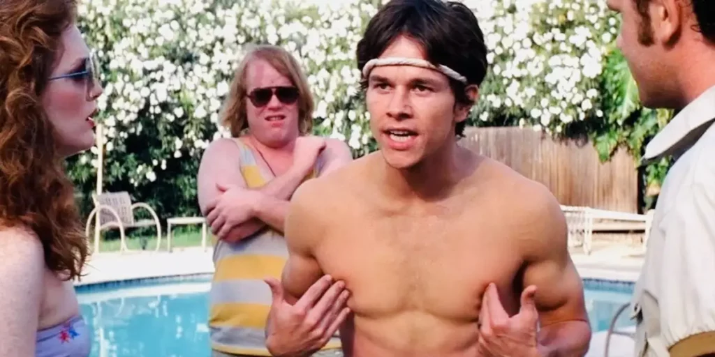 A shirtless man gestures while talking in front of a pool, with another man with sunglasses standing behind him, in the film Boogie Nights