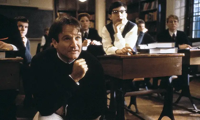 Robin Williams kneels in front of his students, looking ahead, in the film Dead Poets Society, where he played movie teacher John Keating