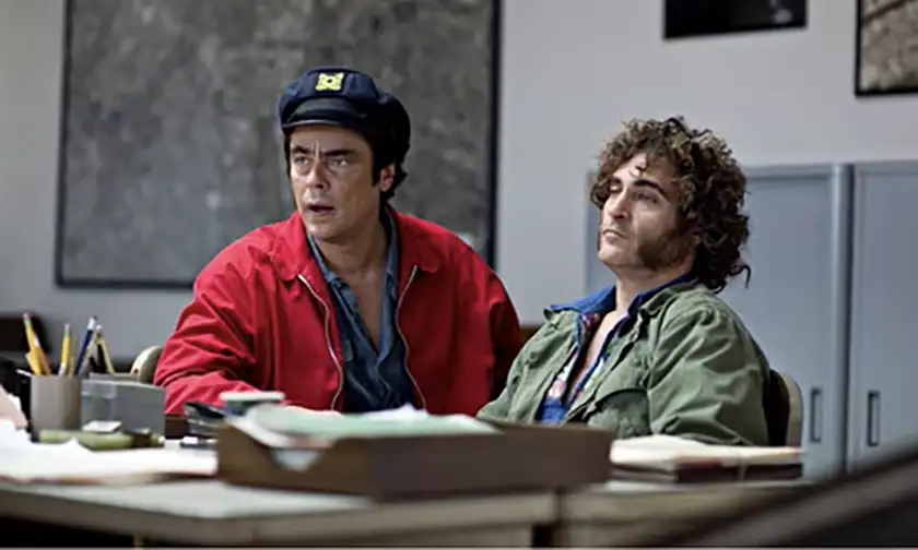 Benicio del Toro and Joaquin Phoenix sit behind a desk in the film Inherent Vice