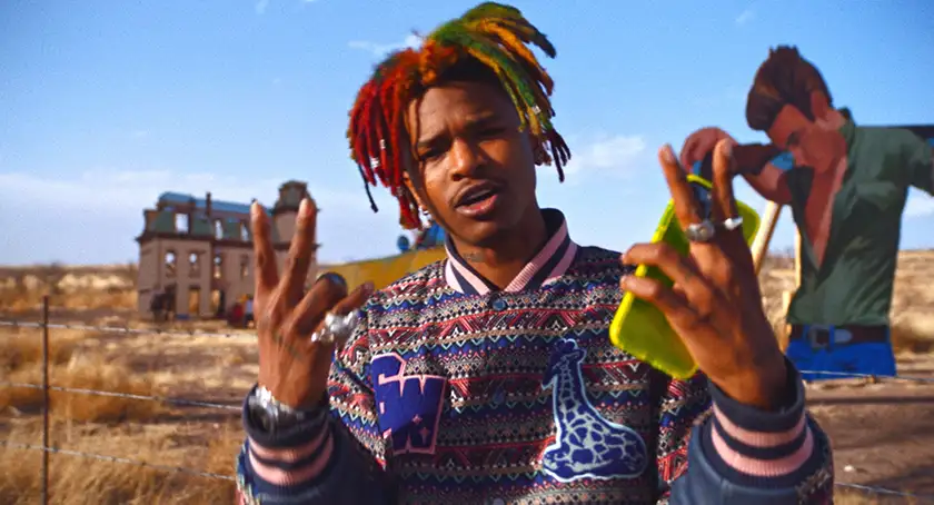 A Black man with multicolored dreadlocks makes peace gestures while singing and holding a brigh yellow iPhone in a still from the movie Lost Soulz
