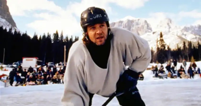 Russell Crowe wears a white sweater and plays ice hockey on a frozen pond in the middle of the mountains, with some people watching behind him, in the movie Mystery, Alaska
