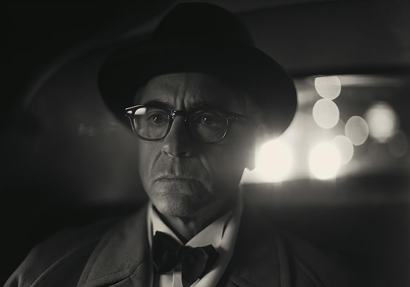 Robert Downey Jr. sits in a car wearing a suit and a hat in a black and white shot from the film, whose line "I Am Become Death" is explained in Loud and Clear Reviews' article