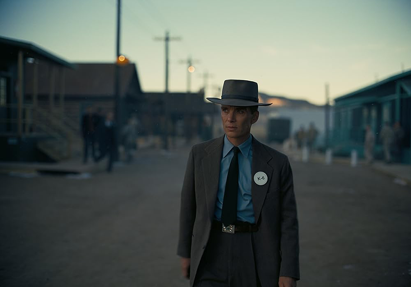 Cillian Murphy stands looking worried in the middle of a road in Los Alamos, wearing a K-6 badge on a suit in the film OPPENHEIMER