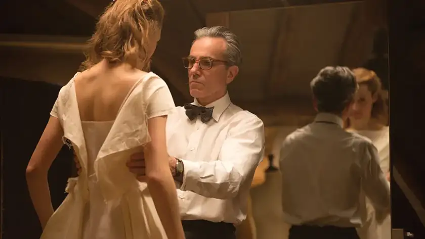 A man adjusts a woman's dress as he is making it, and they both stand in front of a mirror, in the film Phantom Thread