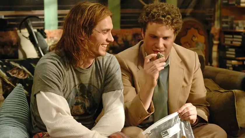 Seth Rogen and James Franco sit on a couch, the former holding weed and the latter smiling and looking at it, in the movie Pineapple Express