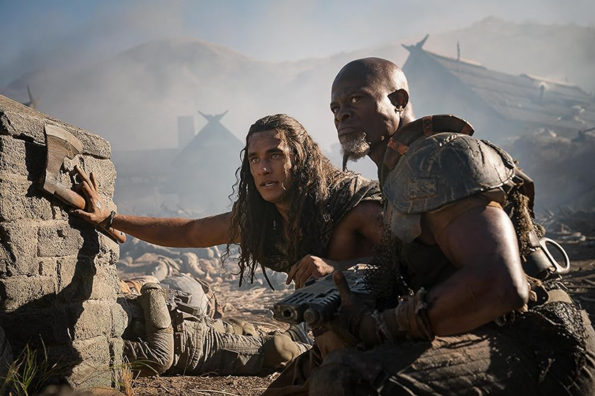 Staz Nair as Tarak and Djimon Hounsou, two men dressed as generals, lean on a stone fence while holding a gun and an axe in Rebel Moon Part Two: The Scargiver