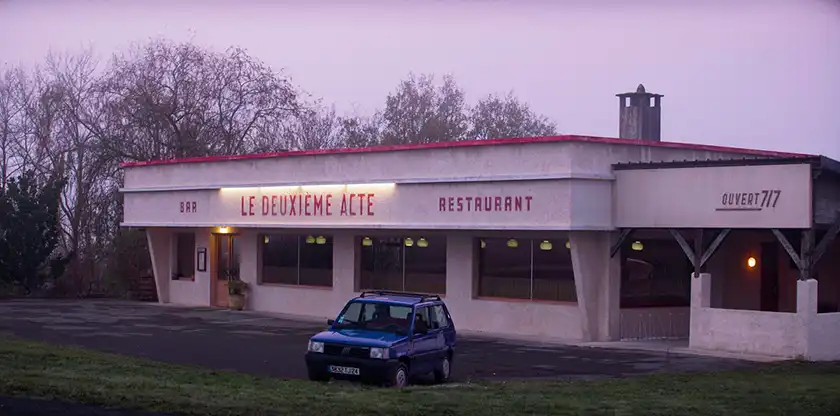 A blue punto is parked in front of a restaurant called Le Deuxieme Acte in The Second Act, one of the 20 films to watch at the 2024 Cannes film festival according to Loud and Clear Reviews