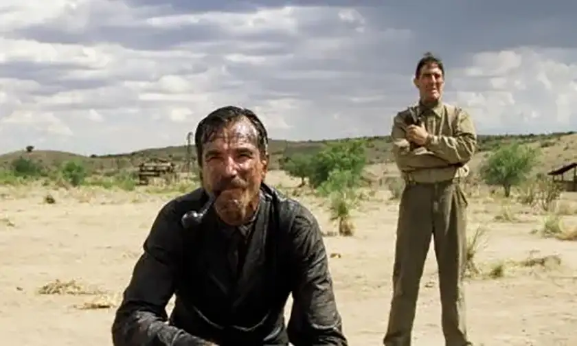 Daniel Day-Lewis crouches in the desert with a pipe in his mouth, a man standing behind him, in the film There Will Be Blood