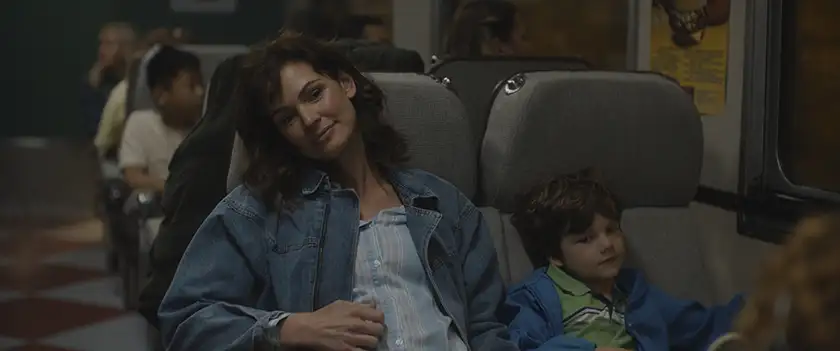 A woman wears a jeans jacket and touches her belly, smiling, while sitting to a train next to a child in the film UNSUNG HERO