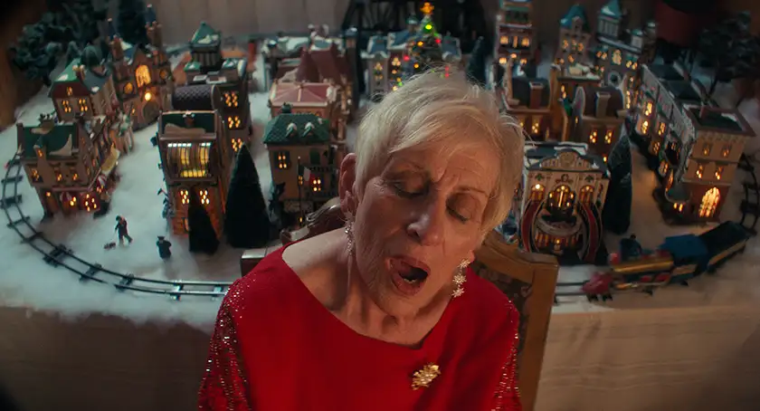 An old woman with a red sweater sings in front of a table, where a toy Christmas town is placed, in the Tyler Taormina film Christmas Eve in Miller's Point, featured in the Loud and Clear interview