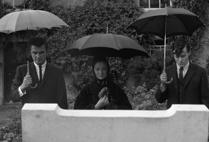 Three people stand looking at a tombstone holding umbrellas in a black and white still from the film Dementia 13