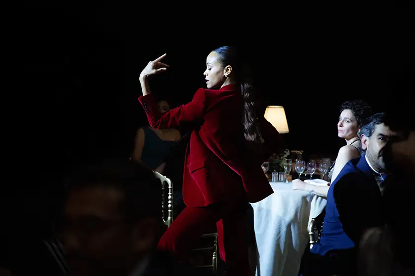 Zoe Saldana wears a red suit and dances in a dark room, with a table behind her, in the film Emilia Pérez