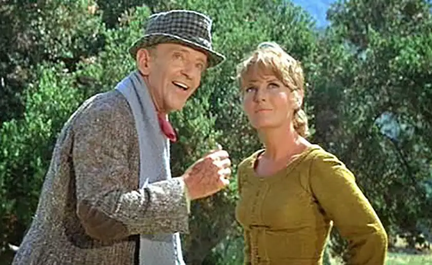 Fred Astaire and Petula Clark stand outdoors surrounded by trees, the former making a gesture and looking at something and the latter looking too, in the film Finian's Rainbow, one of all Francis Ford Coppola movies ranked from worst to best by Loud and Clear Reviews