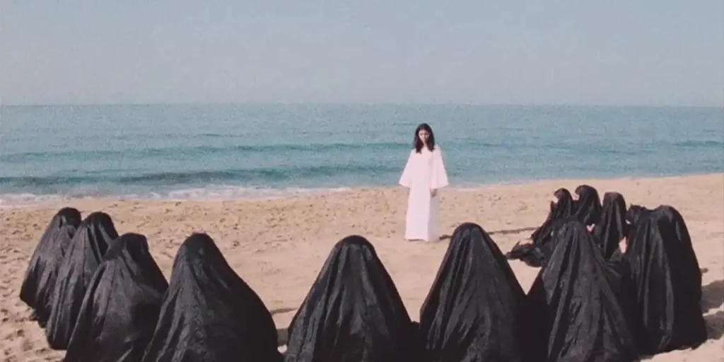 A woman stands on the sand at a beach, with the sea behind her and women dressed in black sitting in a circle around her, in the film Leila and the Wolves