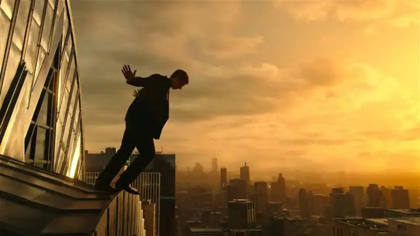 A man stands on the edge of a tall building, about to fall down, with yellow sky behind him, in the film Megalopolis
