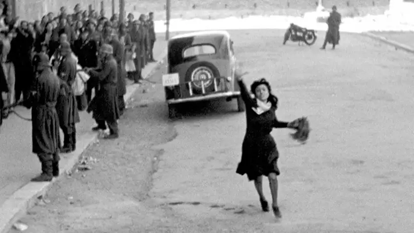 A woman runs raising a hand to ask for help in the middle of a road, with a car behind her and Nazi officers pointing rifles at people on her right, in the film Rome, Open City