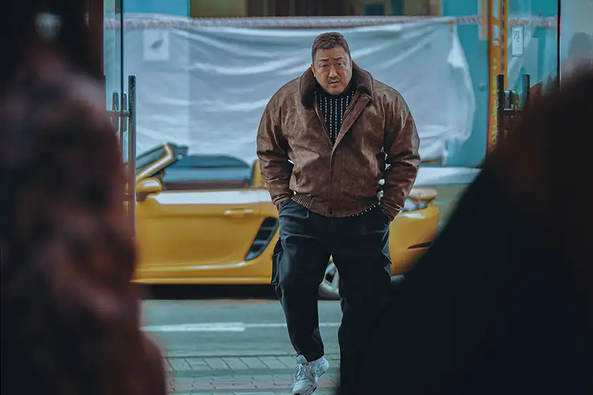 Ma Dong-seok walks with a yellow car behind him in the film The Roundup: Punishment