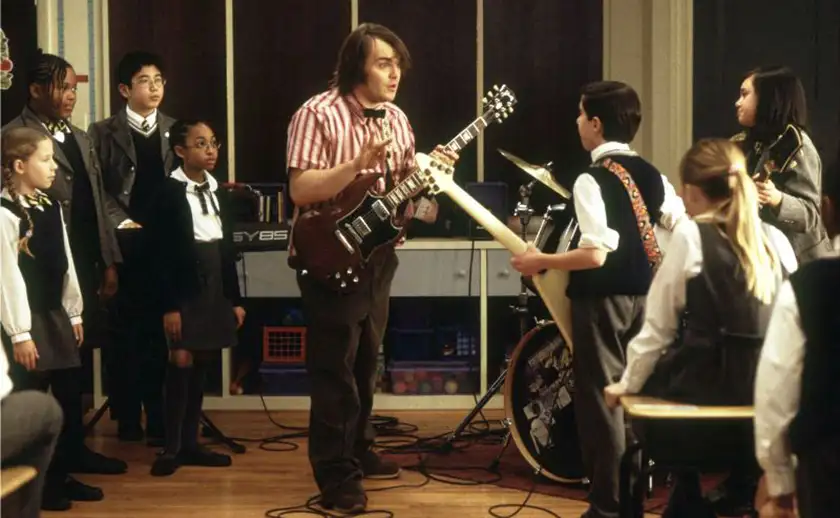 Jack Black holds an electric guitar and teaches children standing around him in the film School of Rock