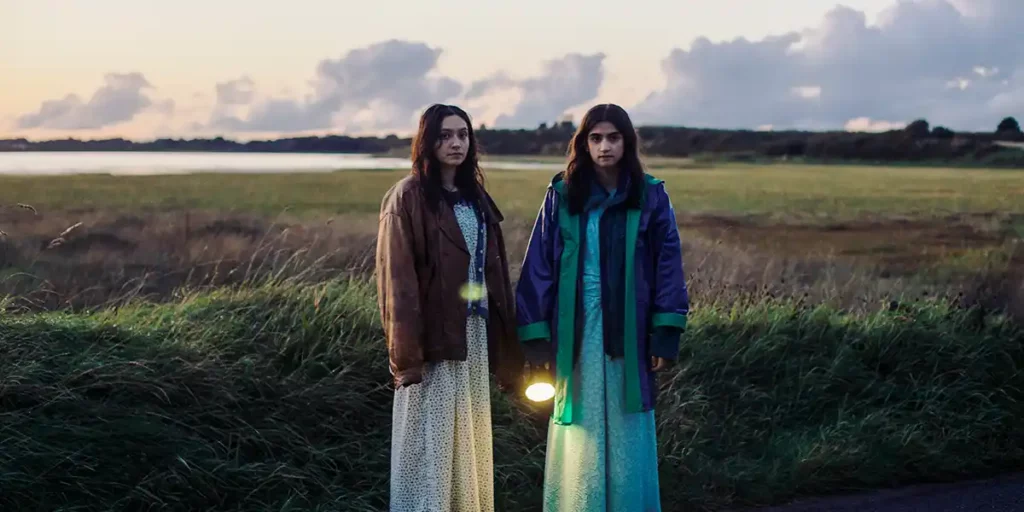 Twi girls stand wearing colorful dresses and holding lanterns in a field at dusk in the film September Says