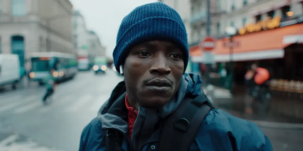 A delivery man looks to his left in the middle of the street in the film The Story of Souleymane