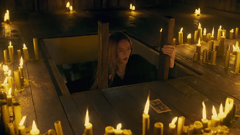 A woman's head emerges from a trapdoor, her face in disbelief as she looks at the lit candles all around her, with a tarot card in front of her